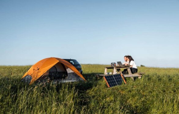 Couple utilisant un ordinateur portable alimenté par un Jackery générateur solaire avec panneau solaire portable près d'une tente orange dans un champ verdoyant