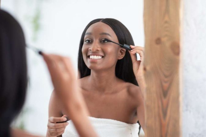 Une jeune femme souriante aux longs cheveux noirs s'applique du mascara sur les cils tout en se regardant dans un miroir pour un maquillage nude.