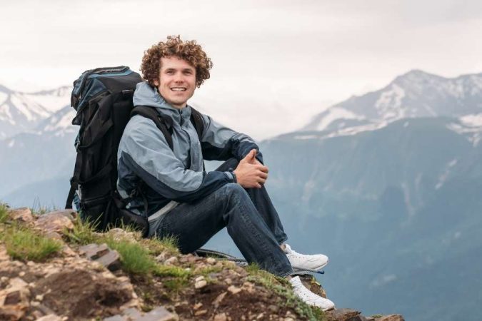 Un jeune homme avec un sac à dos est assis sur un sommet rocheux devant des montagnes enneigées.