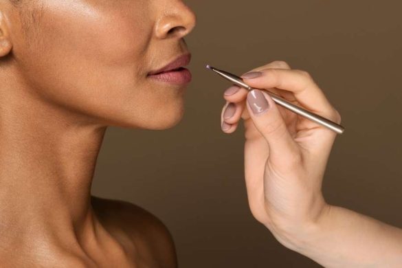 Gros plan sur une main qui utilise un pinceau fin pour appliquer du rouge à lèvres aux teintes naturelles sur la bouche d'une femme.